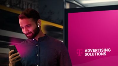 Man looking at mobile phone, standing in front of outdoor digital display with “T-Mobile Advertising Solutions” on it.