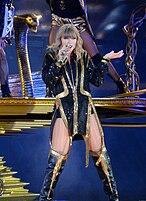 Taylor Swift singing on a microphone, dressed in a black suit with golden accents