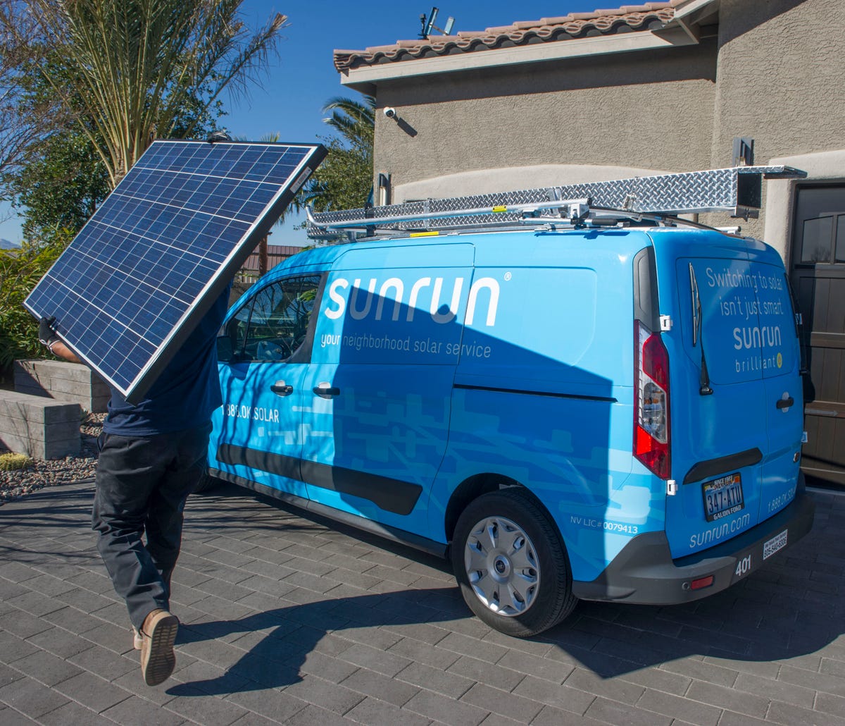 A person carries a solar panel past a blue Sunrun van.