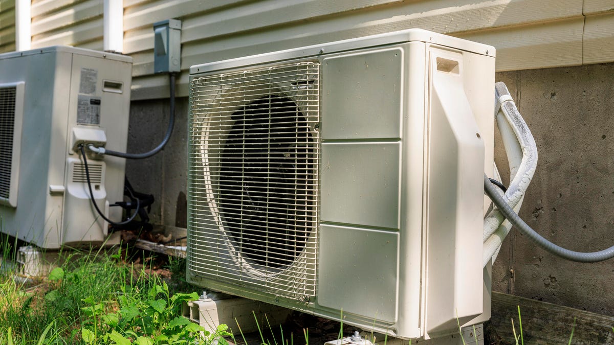 A heat pump sitting outside a house with grass and weeds in front of it.