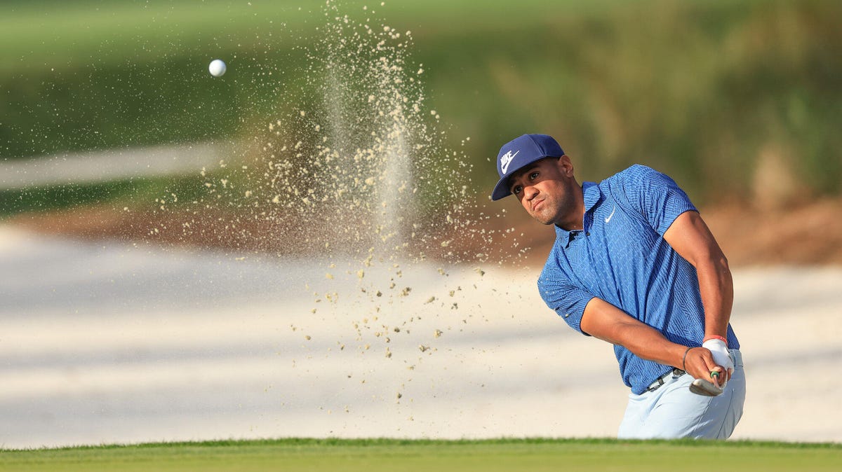 Golfer Tony Finau playing a shot from a bunker, rising sand in the air.