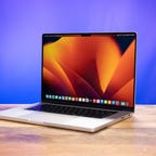 The Apple MacBook Pro 16 sitting on a wood desk, open and angled to the left showing the orange-flower desktop, against a purple-blue background