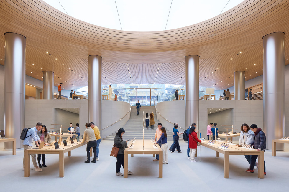 Customers look at an array of Apple products on long tables as well as walk up the store’s central staircase.