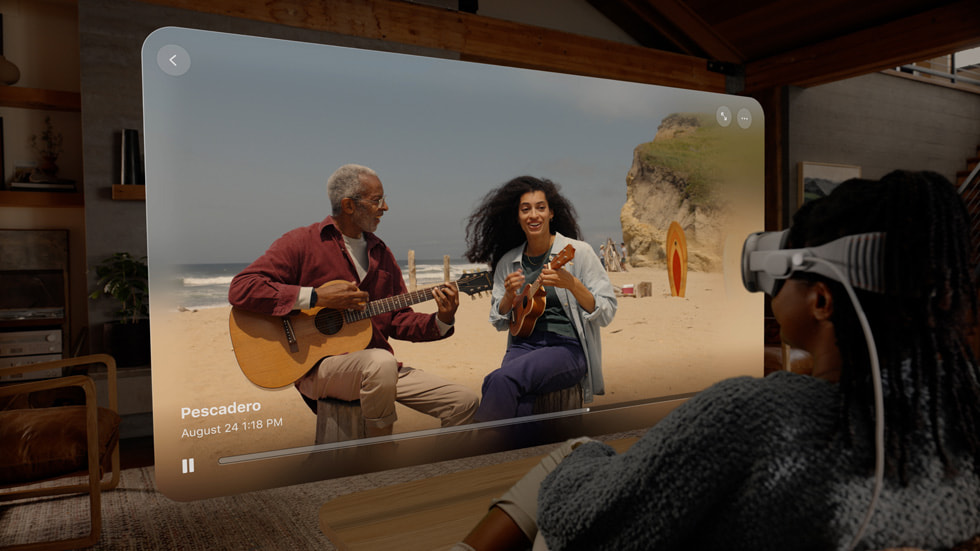 Una persona che indossa un Apple Vision Pro per guardare un video spaziale di due persone che suonano la chitarra su una spiaggia, mostrato in una finestra nel suo salotto.