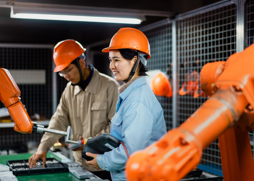 Two supply chain workers in hard hats use equipment in a factory setting.