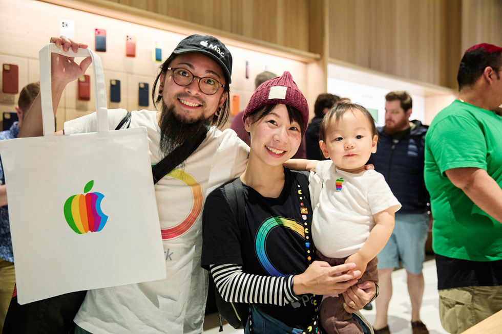 Casal de clientes com camisetas da Apple. Ela segura um bebê e ele, uma sacola da Apple.