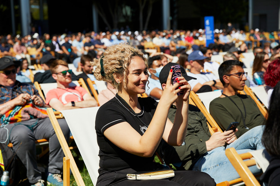 Uczestnicy siedzący przed kafeterią Caffè Macs na kampusie Apple Park oglądają prezentację, która otwiera konferencję WWDC22.