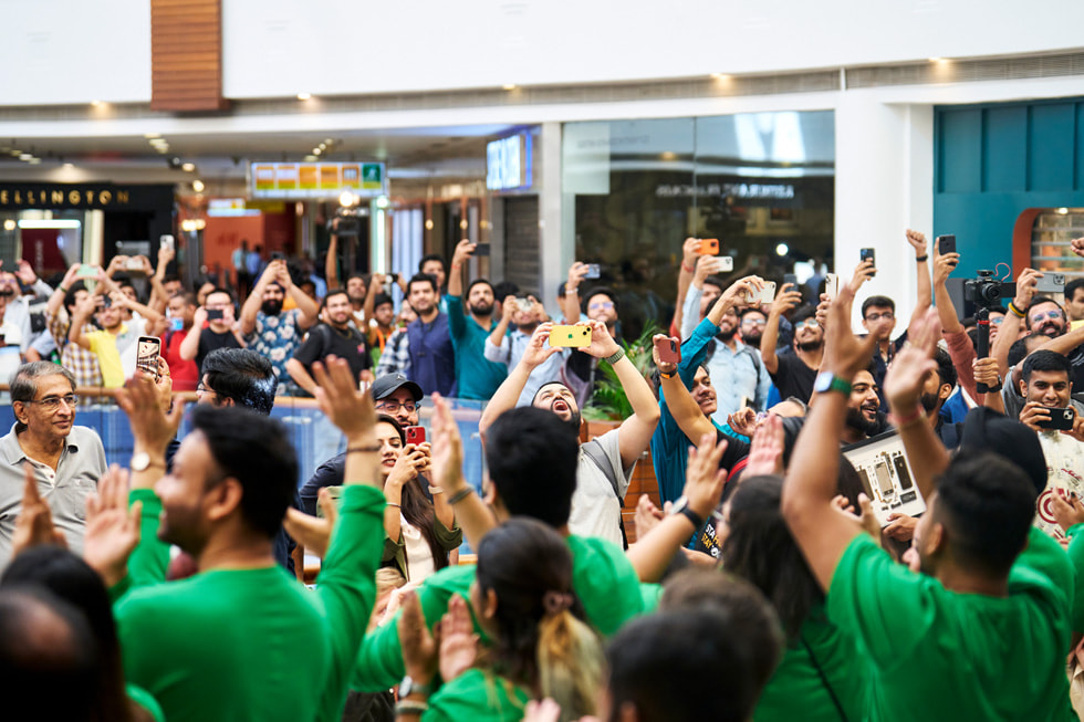 Un grand groupe de personnes attend devant Apple Saket, plusieurs lèvent les bras pour prendre des photos.