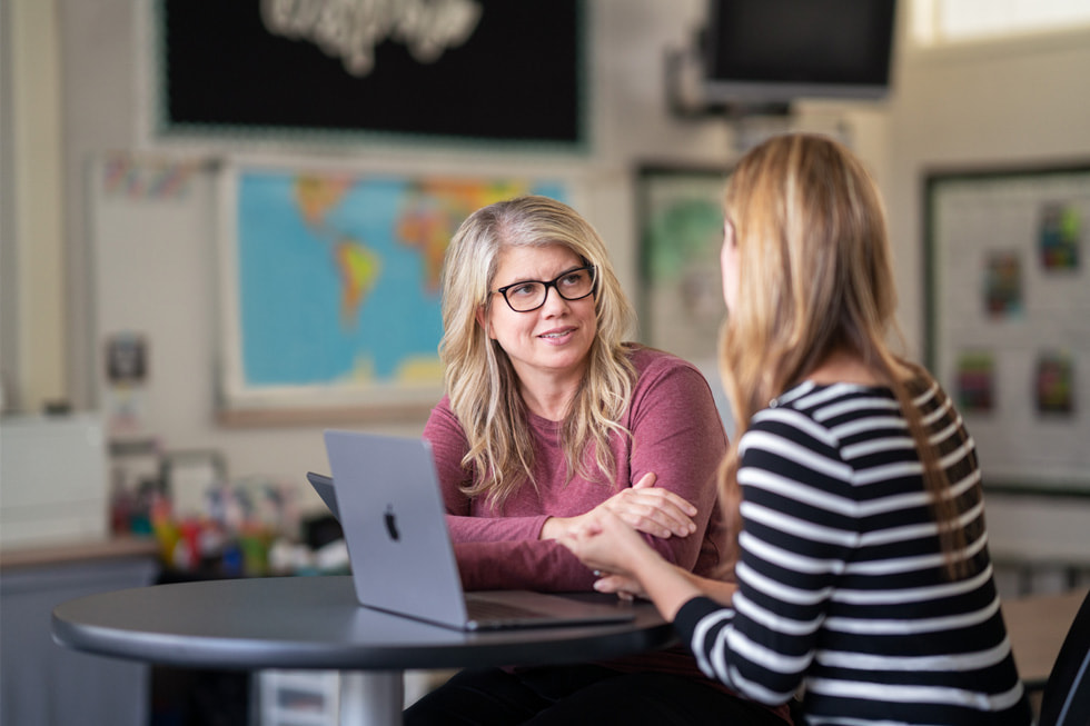 Marnie Luevano, Lehrcoach für den Einsatz von Technologie im Unterricht bei Downey Unified, spricht mit einem anderen Erwachsenen in einem Klassenzimmer mit einem MacBook Air auf dem Tisch.