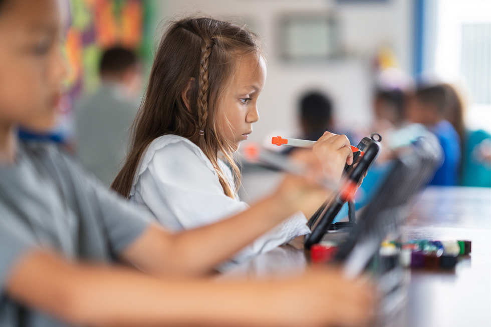 A young student holds a stylus up to iPad in a classroom setting.