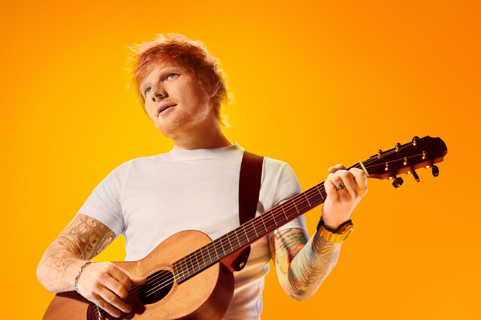 O cantor e compositor Ed Sheeran toca violão em frente a um fundo laranja.