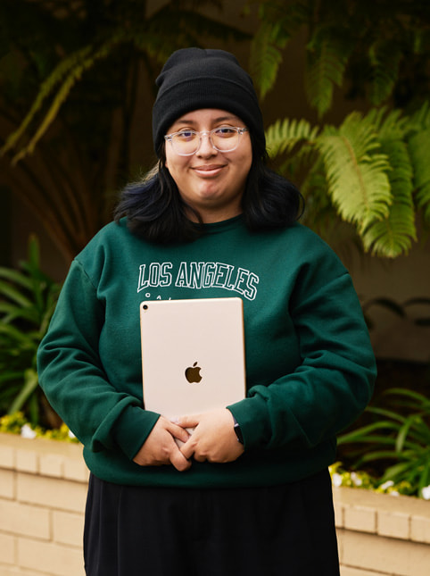 Un retrato de Angela Ibarra, estudiante de Exceptional Minds. Angela lleva un suéter verde que dice "Los Angeles, California", anteojos de marco transparente y un gorro negro.