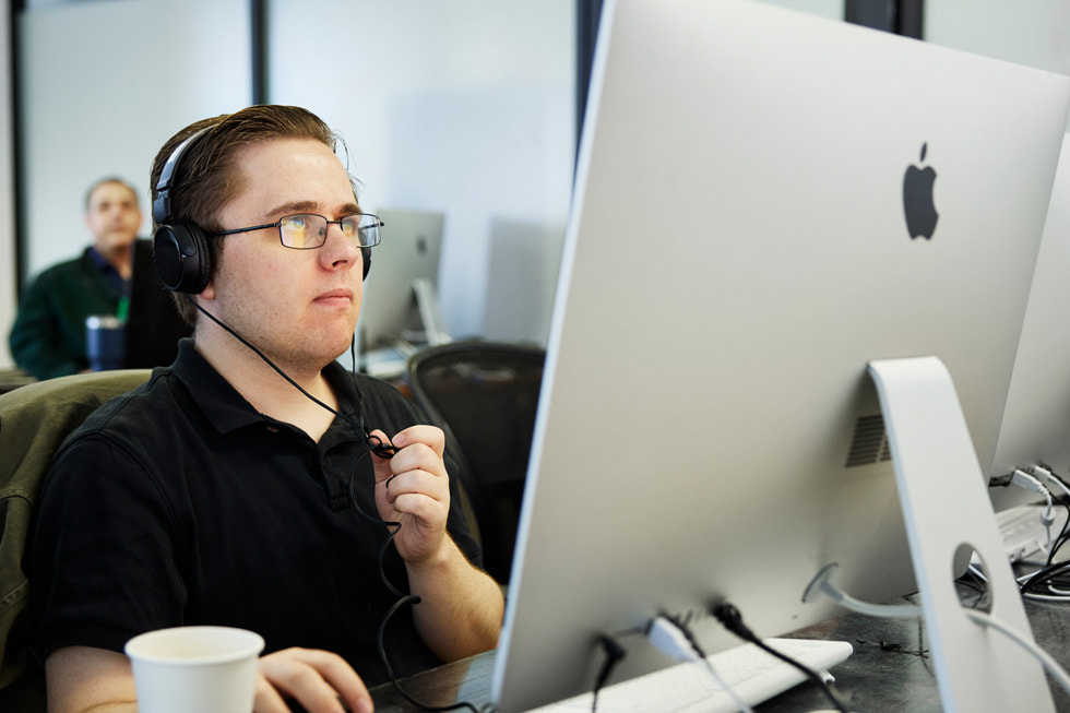 Der Exceptional Minds-Student Matthew Rohde bei der Arbeit an einem Mac in einem Klassenzimmer auf dem Campus. Rohde trägt ein schwarzes Poloshirt und Kopfhörer.