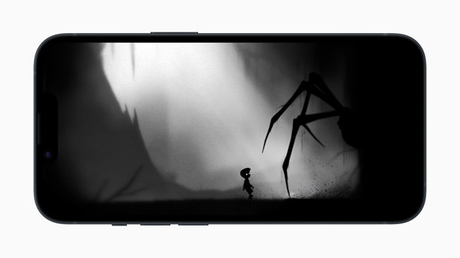 På iPhone 14 vises et stillbillede fra spillet Playdeads LIMBO+, hvor en kæmpeedderkop lurer i skyggerne nær et lille barn.