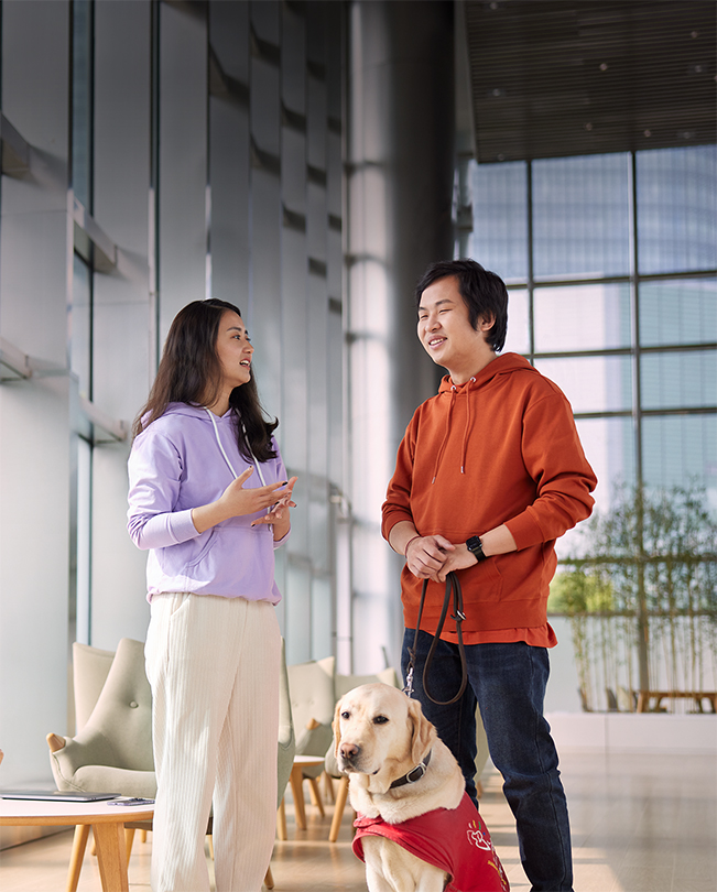 Dwoje pracowników Apple rozmawiających w jasno oświetlonym miejscu, spośród których jeden trzyma na smyczy psa opiekuna.
