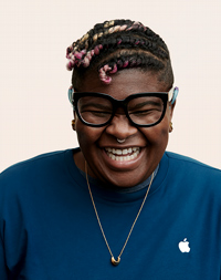 Apple Retail Mitarbeiterin mit Kopftuch und Brille lächelt in die Kamera. 