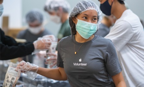 Stażystka Apple w koszulce z logo wolontariatu Apple. Uśmiecha się i patrzy wzrokiem poza kadr, pakując rzeczy w ramach akcji wolontariackiej.