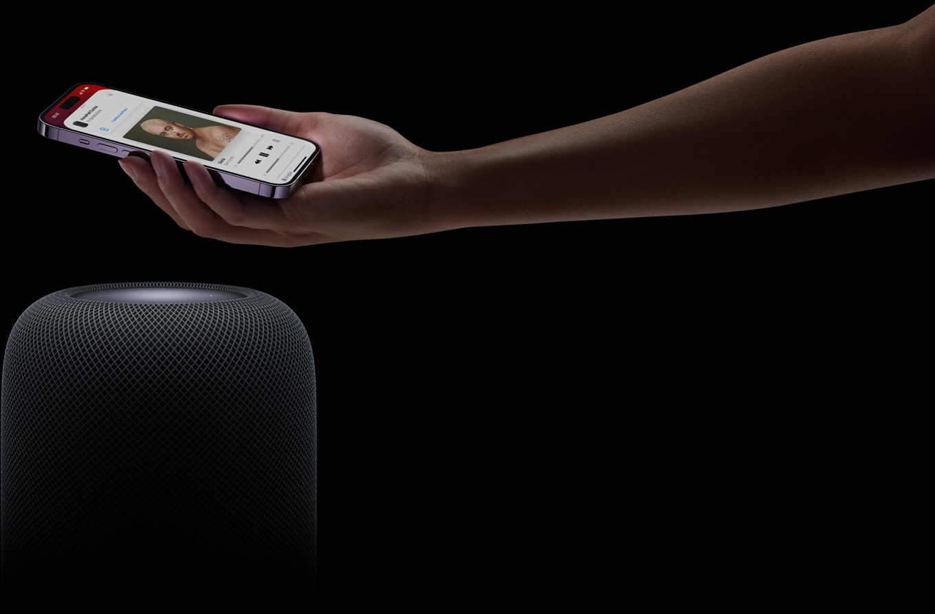 Un braccio che entra nell’immagine da destra, con un iPhone tenuto nel palmo della mano sopra un HomePod