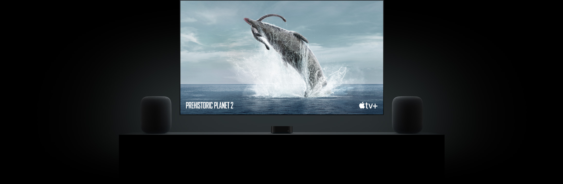 Suuri taulu-TV, jossa näkyy upea kuva dinosauruksesta Prehistoric Planet -ohjelmasta. TV on asennettu Apple TV:n yläpuolelle, ja sen sivuilla on kaksi HomePod-kaiutinta olohuoneen senkin päällä