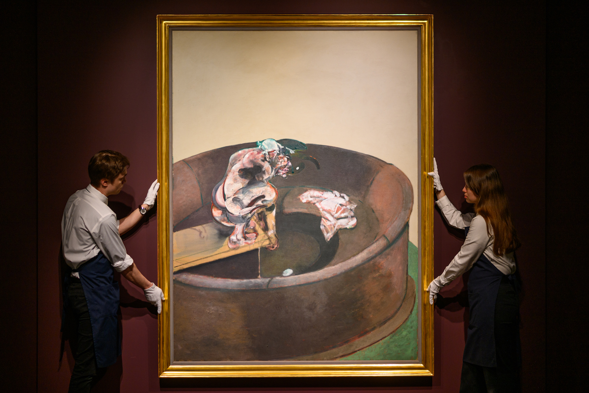 Two aproned auction house workers hold up a painting of a crouching man on a diving board. His face and body are abstracted.