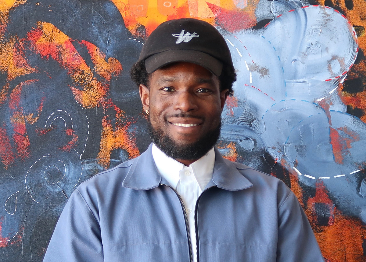 A portrait of Walter Price, a Black man wearing a black baseball hat, powder blue jacket, and white collar shirt. He stands before an abstract painting seen in detail.