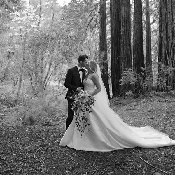 Bride in Strapless Wedding Dress Holding Bouquet Kissing Groom in Black Tuxedo in Wooded Area
