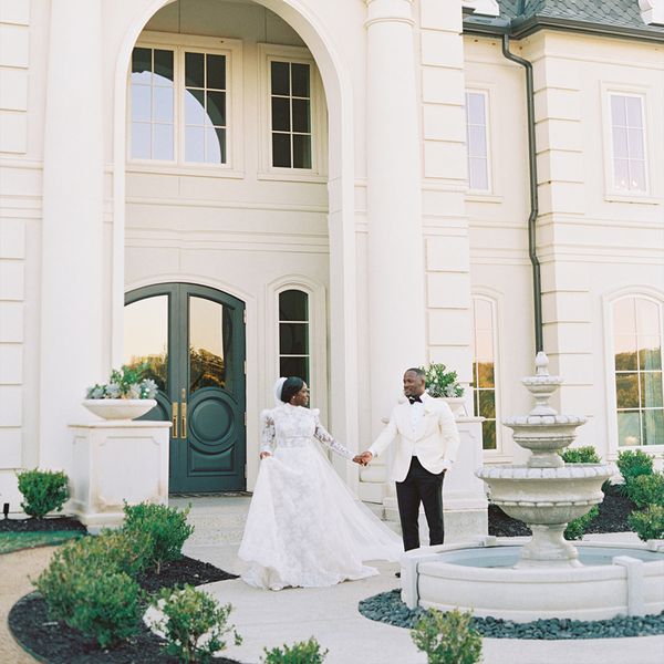 Bride in Long-Sleeved Wedding Dress and Groom in White Tuxedo Jacket Holding Hands in Front of White Mansion With Green Doors