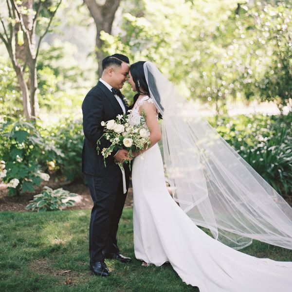 bride holding lush bouquet and groom in black tuxedo pose in garden during wedding first look