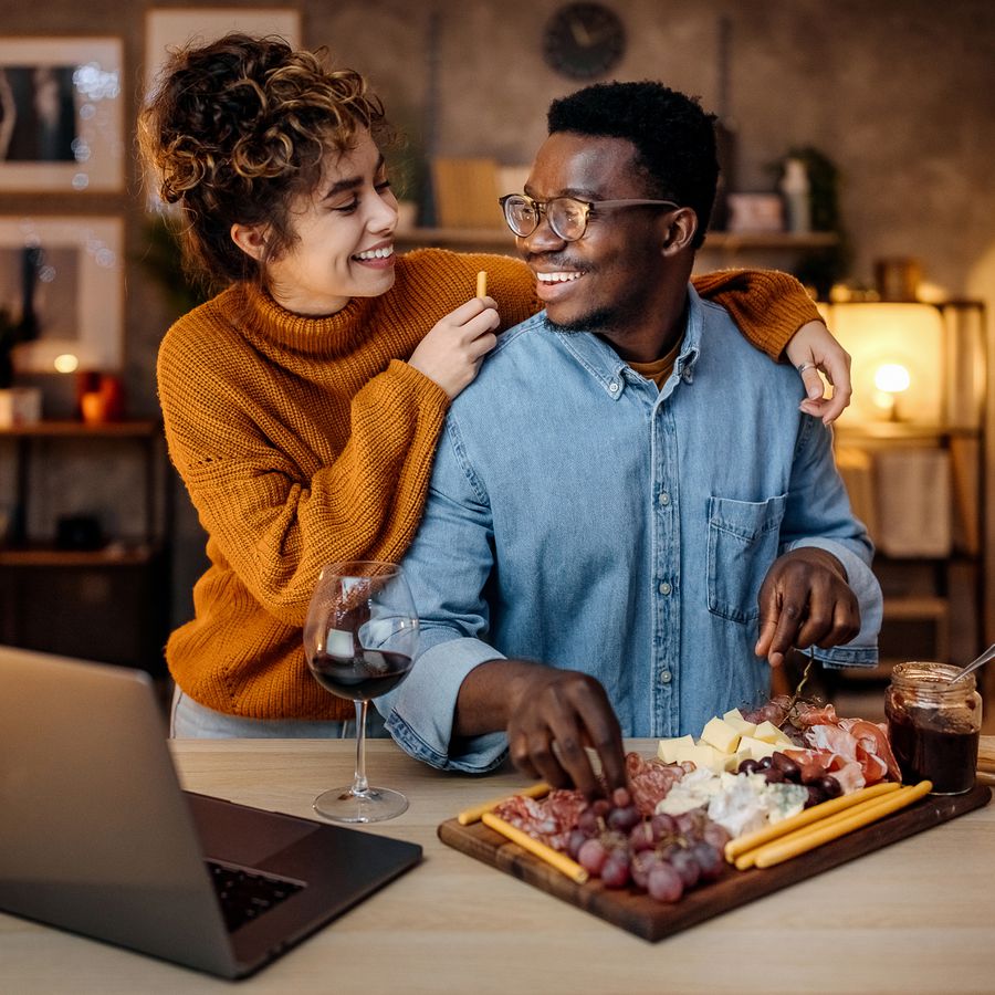 Young couple smiling while making charcuterie board and drinking wine during date night indoors at home.
