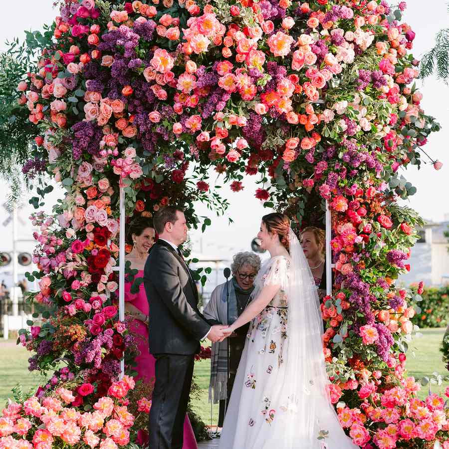 Bride and groom holding hands in front of a peach, pink, and purple floral arch