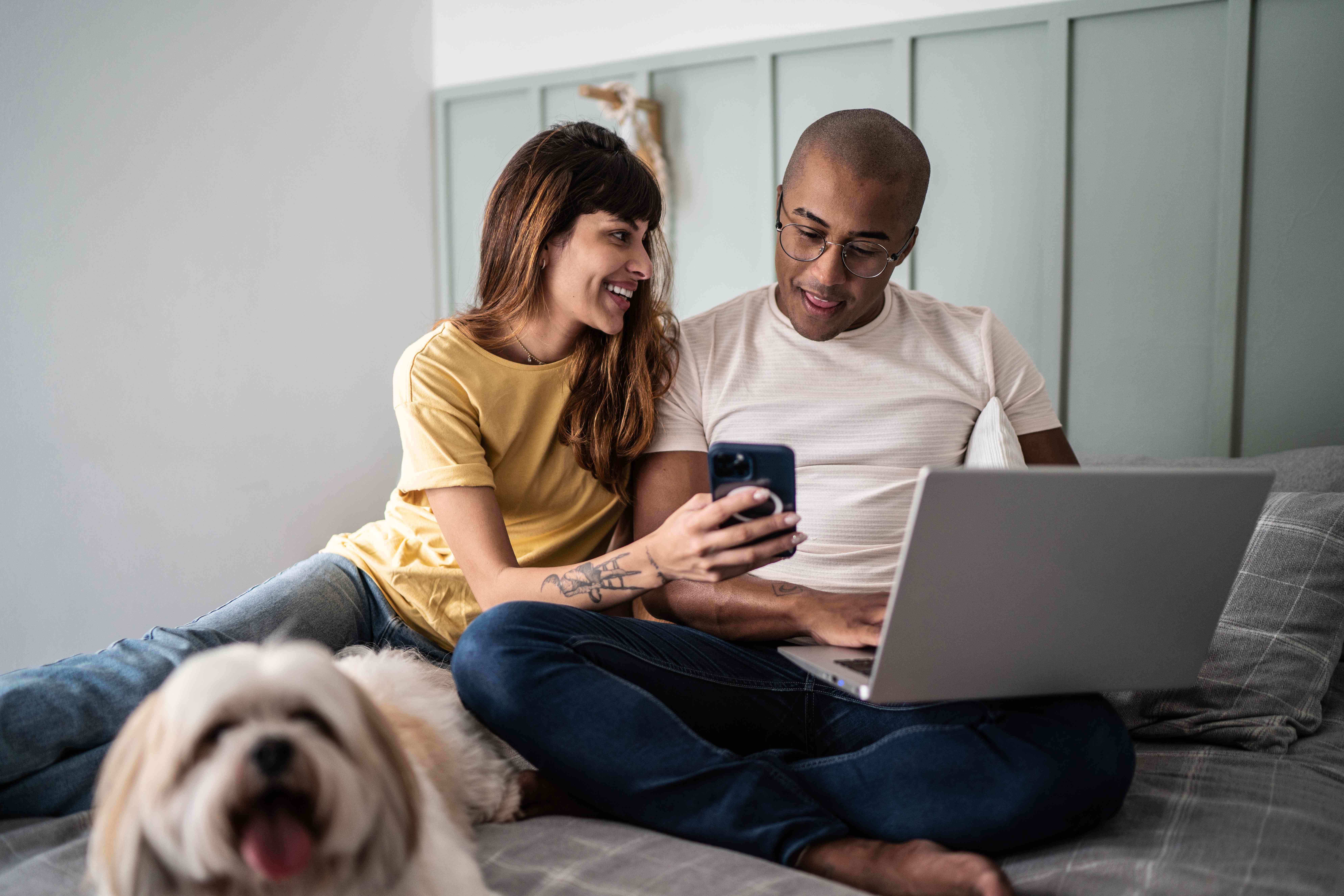 Young woman smiling as she shows something to her male partner on her phone, while sitting on the bed with a dog