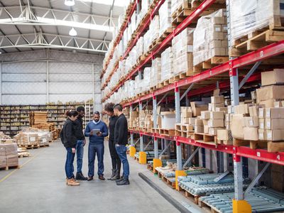 Group of people in a warehouse surrounding by boxes all looking at a tablet held by one person