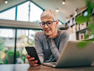 Older woman looking happily at her smartphone with a laptop also open in front of her