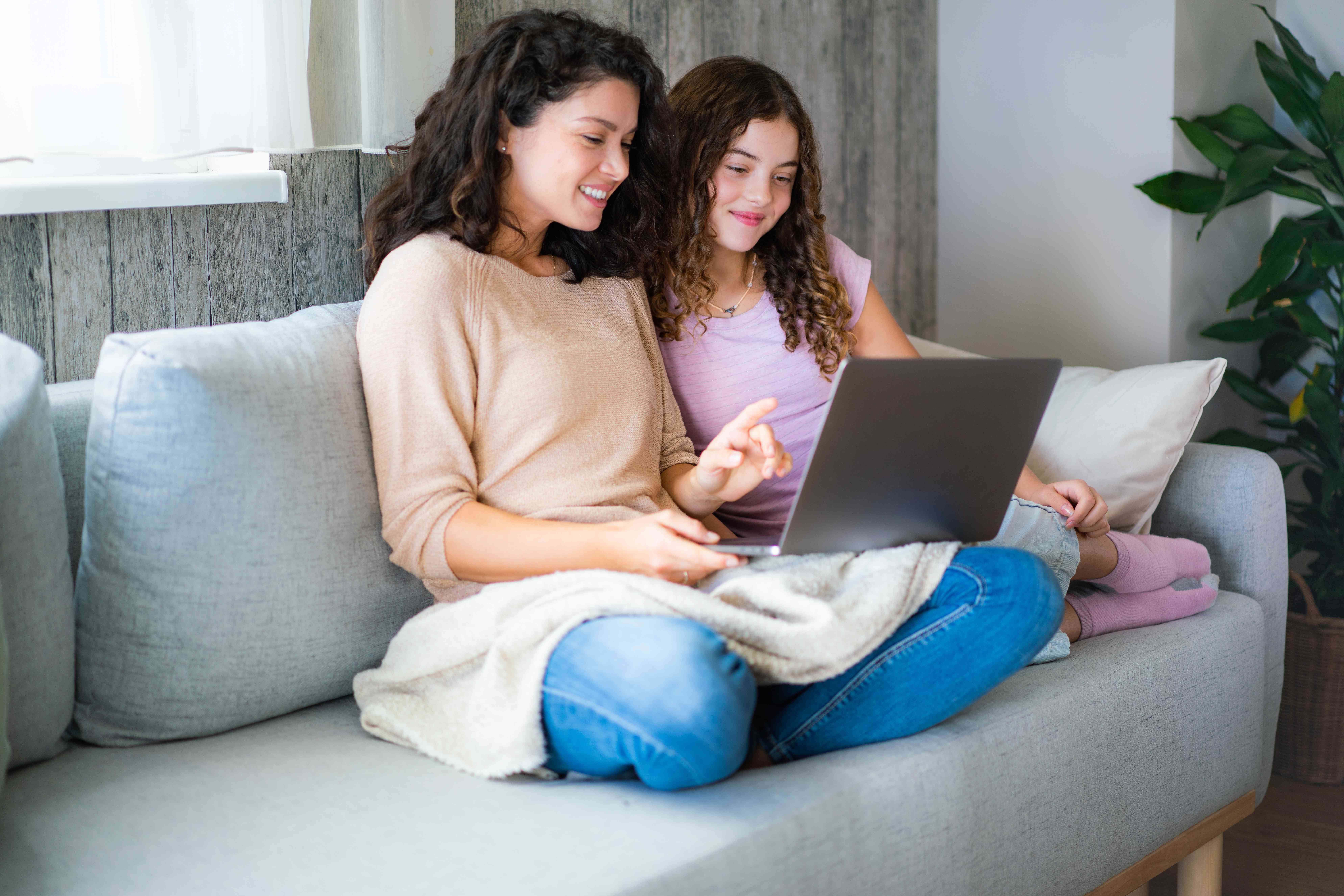 Mom and young daughter at home on couch, looking together at laptop