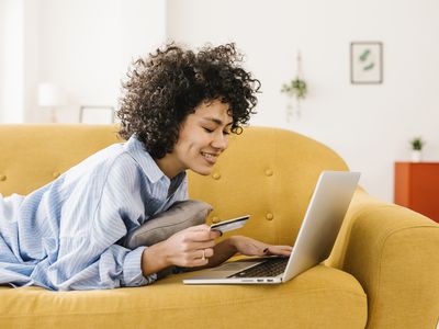 A woman uses a laptop with a credit card on the couch.