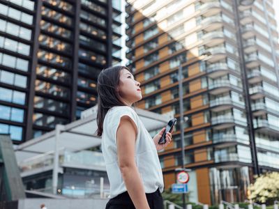 Young woman with a smartphone stands in front of a skyrise building. 