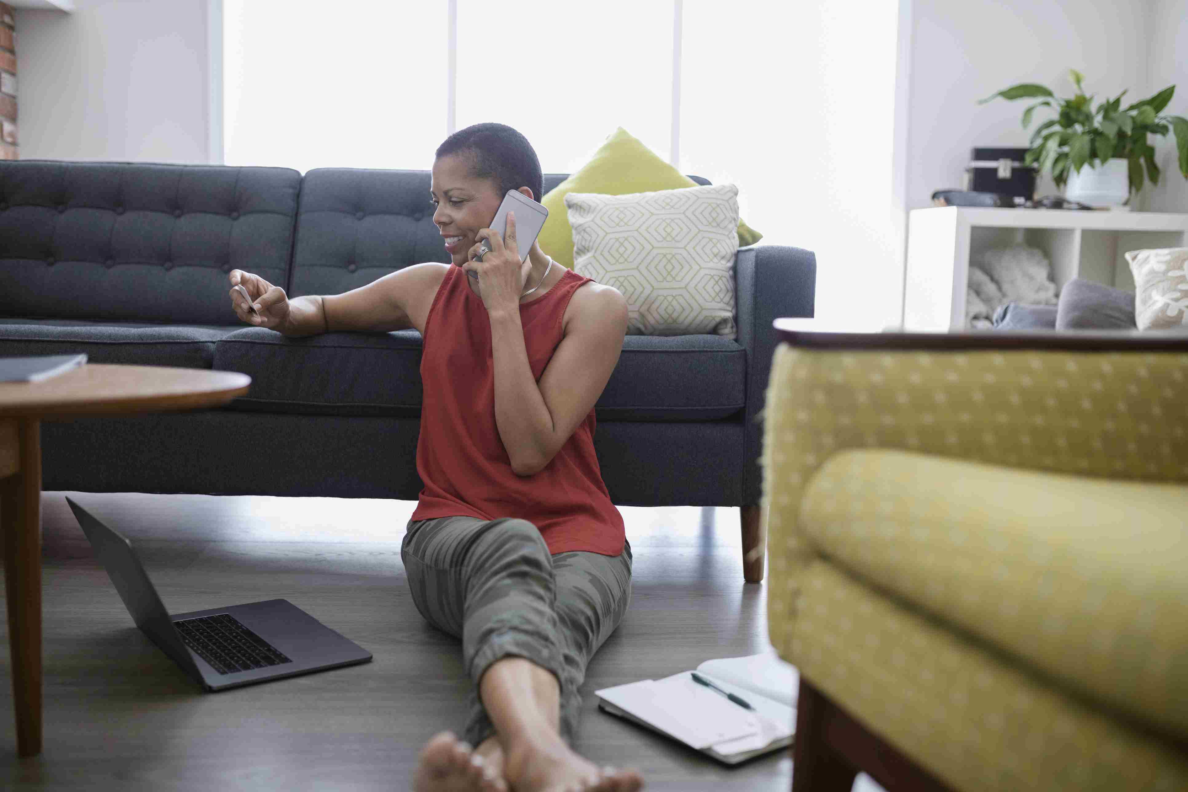 Mature woman with credit card talking on cell phone, using credit card at laptop on living room sofa