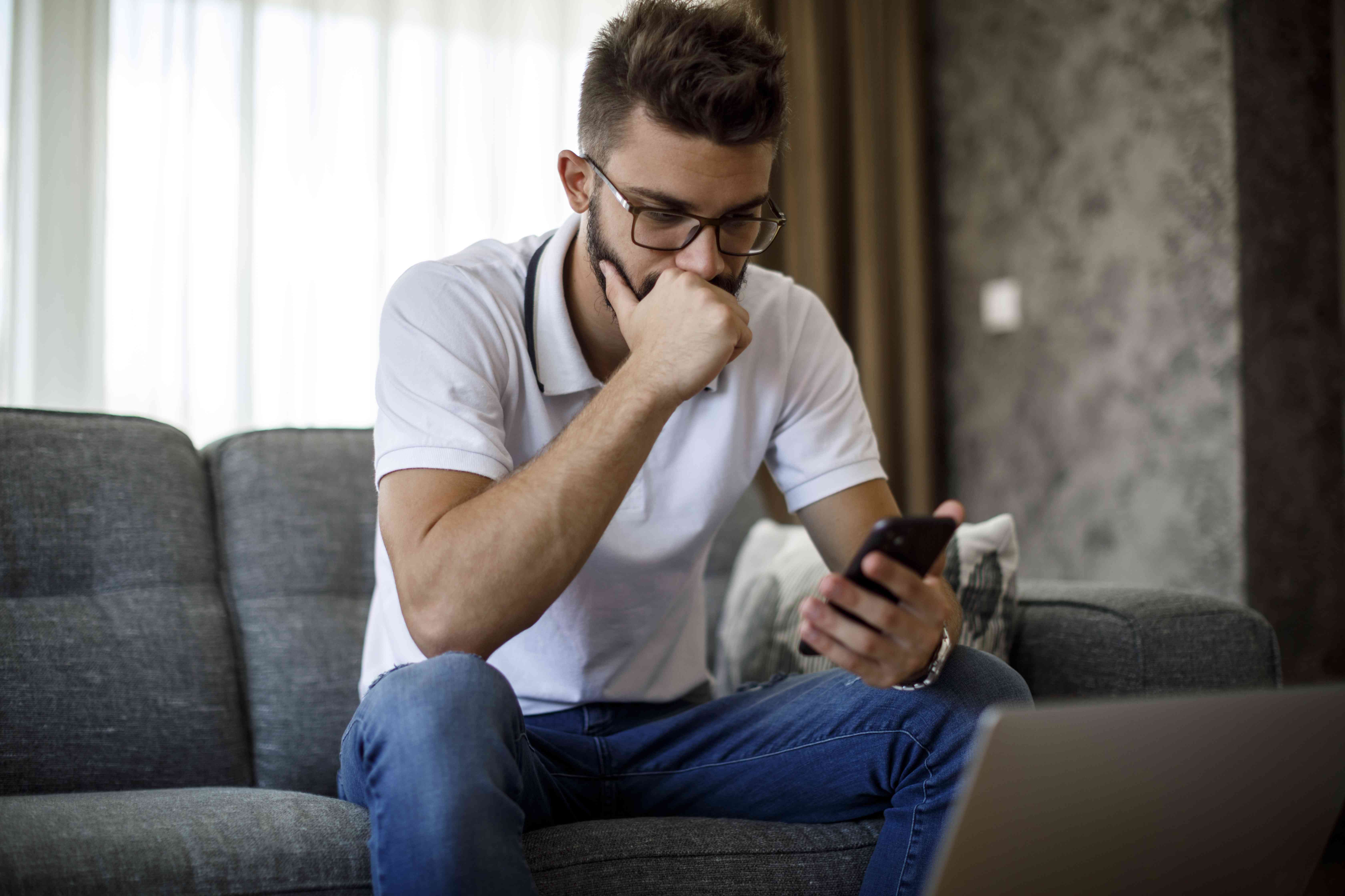 Young man sitting on living room couch and looking intently at his smartphone