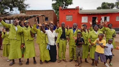 A group of people (women, men and children) of an African cooperative is waving their hands to the camera, standing in front of a red brick house.