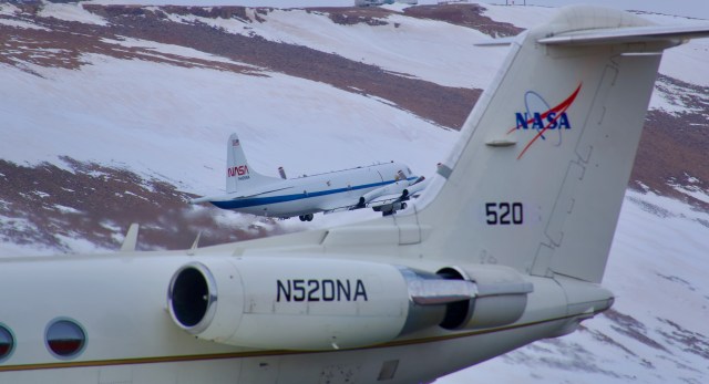 An image of two aircraft in front of a hill covered in snow and rock. In the foreground is the tail end of a white jet, filling the bottom and right side. The NASA logo and number 520 are on the tail. Behind the jet, in the middle of the image, another white aircraft takes off. It’s white with a blue horizontal stripe, with the NASA ‘worm’ logo on the tail. The brown and white hillside fills the rest of the frame.