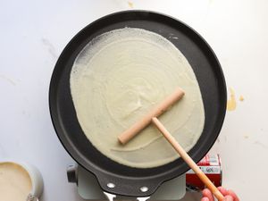 A person using a spreader to spread crepe batter onto a crepe pan.
