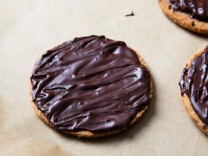 A vegan chocolate-covered digestive biscuit resting on parchment paper. There are two other biscuits on the right periphery of the image.