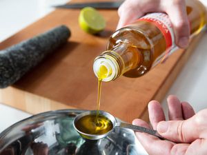 Mustard oil being poured into a measuring spoon. A cutting board, knife, pestle, and half a lime are out of focus in the background.