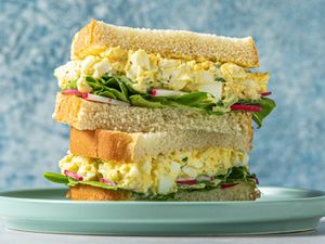 An egg salad sandwich, cut in half, with the two halves stacked on top of each other. The sandwich contains egg salad, butter lettuce, and thinly sliced radishes.