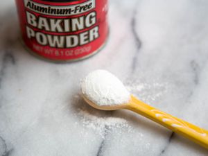 A container of baking powder next to a spoon full of baking powder. 