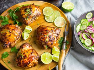 Four Vietnamese baked chicken thighs, served on a wooden serving platter with cut limes and a bowl of cucumber salad.