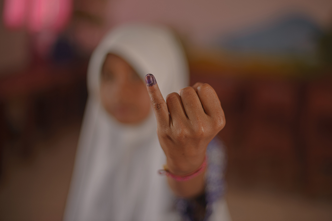 The image features a person wearing a white hijab, showing their index finger with purple ink on it, indicating that they have just vaccinated.