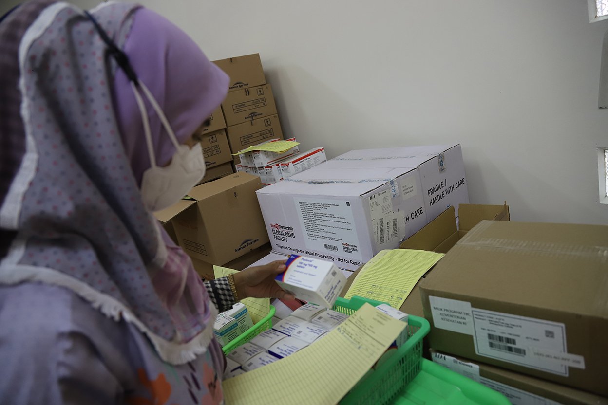 The image shows an individual in a hijab examining stacked boxes of medication. They are holding and looking at a package of medicine, seemingly conducting inventory or stock checking in a storage room or pharmacy.