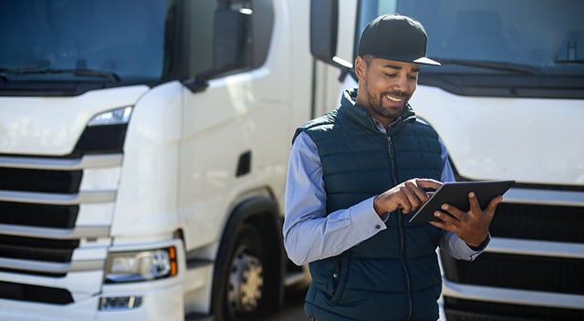 A truck driver uses a tablet in front of two parked semi-trucks.