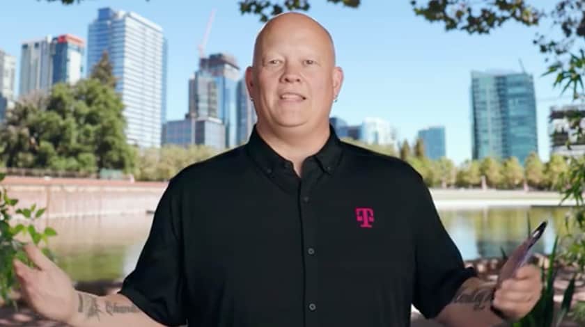 Smiling man wears a black shirt with the T-Mobile logo on it and stands with his arms extended